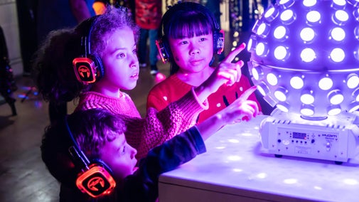 Three children enjoing the silent disco at Plas Newydd, Anglesey. They're wearing lit up headphones and are clustered around a lit up disco ball.